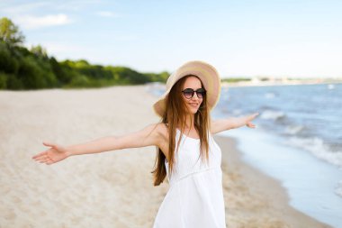 Okyanus sahilinde mutlu, mutlu, mutlu bir kadın şapkası, güneş gözlüğü ve açık elleriyle duruyor. Beyaz yaz elbisesi içinde çok kültürlü bir kadın modelin portresi seyahat sırasında doğanın tadını çıkarıyor.