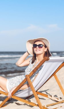 Mutlu esmer kadın güneş gözlüğü takıyor ve şapka takıyor. Okyanus sahilinde tahta bir iskemlede dinleniyor..