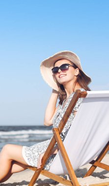 Mutlu esmer kadın güneş gözlüğü takıyor ve şapka takıyor. Okyanus sahilinde tahta bir iskemlede dinleniyor..