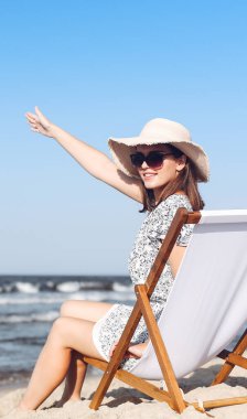 Mutlu esmer kadın, okyanus sahilinde ahşap bir iskemlede oturmuş el sallıyor ve biriyle selamlaşıyor..