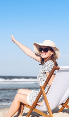 Mutlu esmer kadın, okyanus sahilinde ahşap bir iskemlede oturmuş el sallıyor ve biriyle selamlaşıyor..