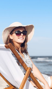 Mutlu esmer kadın güneş gözlüğü takıyor ve şapka takıyor. Okyanus sahilinde tahta bir iskemlede dinleniyor..