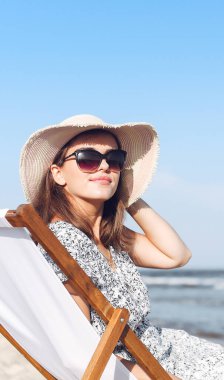 Mutlu esmer kadın güneş gözlüğü takıyor ve şapka takıyor. Okyanus sahilinde tahta bir iskemlede dinleniyor..