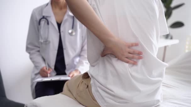 医師と患者さんは診療所にいます 女性が背中をこすっている間 メモを作るためにクリップボードを使用して白いコートの整形外科 閉じます 理学療法の概念 — ストック動画