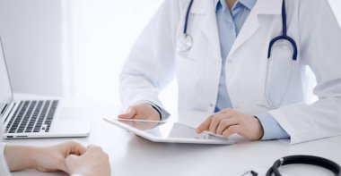 Doktor ve hasta klinikteki masada karşılıklı oturuyorlar. Tablet bilgisayar kullanan kadın doktorların ellerine odaklan, kapat. Tıp konsepti.
