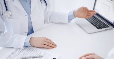 Doktor ve hasta klinikteki masada karşılıklı oturuyorlar. Kadın doktorların elleri dizüstü bilgisayarın monitörünü işaret ediyor, yakın çekim. Tıp konsepti.