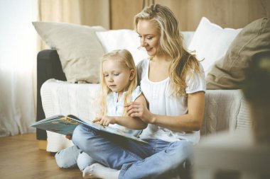 Mutlu bir aile. Sarışın genç anne ahşap zeminde otururken sevimli kızına kitap okuyor. Annelik kavramı.