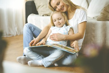 Mutlu bir aile. Sarışın genç anne ahşap zeminde otururken sevimli kızına kitap okuyor. Annelik kavramı.