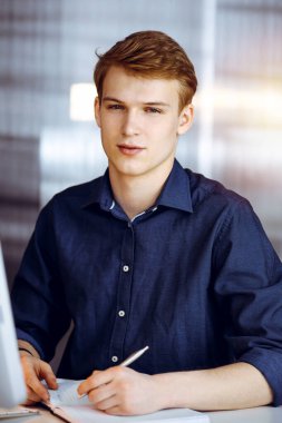 Genç sarışın işadamı karanlık bir ofiste bilgisayarla çalışıyor, arka planda ışık parıltısı var..