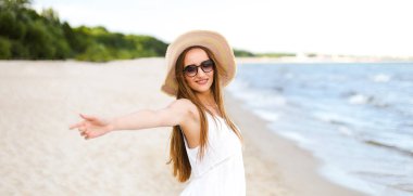 Okyanus sahilinde mutlu, mutlu, mutlu bir kadın şapkası, güneş gözlüğü ve açık elleriyle duruyor. Beyaz yaz elbisesi içinde çok kültürlü bir kadın modelin portresi seyahat sırasında doğanın tadını çıkarıyor.