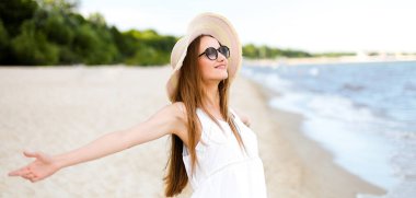 Okyanus sahilinde mutlu, mutlu, mutlu bir kadın şapkası, güneş gözlüğü ve açık elleriyle duruyor. Beyaz yaz elbisesi içinde çok kültürlü bir kadın modelin portresi seyahat sırasında doğanın tadını çıkarıyor.