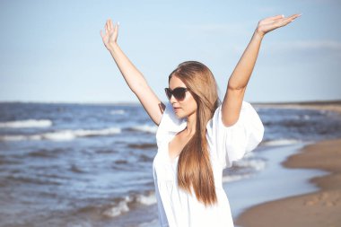 Mutlu sarışın kadın okyanus sahilinde beyaz bir elbise ve güneş gözlüğü ile el kaldırıyor..