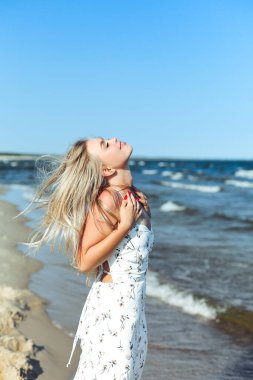 Mutlu sarışın kadın okyanus sahilinde mutlu mesut ayakta duruyor. Beyaz yaz elbisesi içinde bir kadın modelin portresi açık havada tatil sırasında doğanın tadını çıkarıyor..