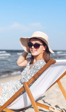 Mutlu esmer kadın güneş gözlüğü takıyor ve şapka takıyor. Okyanus sahilinde tahta bir iskemlede dinleniyor..