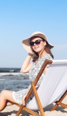 Mutlu esmer kadın güneş gözlüğü takıyor ve şapka takıyor. Okyanus sahilinde tahta bir iskemlede dinleniyor..