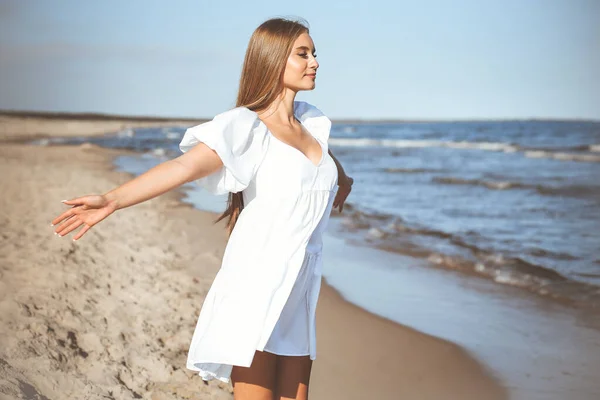 Happy, beautiful woman on the ocean beach standing in a white summer dress, open arms.
