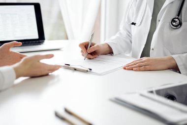 Doktor ve hasta klinikteki beyaz masada oturup bir şeyler tartışıyorlar. Kadın hekim bir kayıt formunu doldururken dinliyor. Tıp konsepti.