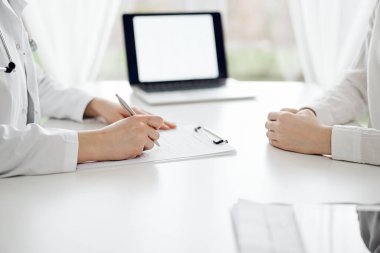Doktor ve hasta klinikteki beyaz masada yan yana oturuyorlar. Kadın hekim bir kayıt formunu doldururken dinliyor. Tıp konsepti.