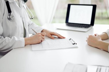 Doktor ve hasta klinikteki beyaz masada yan yana oturuyorlar. Kadın hekim bir kayıt formunu doldururken dinliyor. Tıp konsepti.
