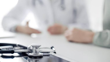 Doktor ve hasta klinikteki masada oturmuş bir şey tartışıyorlar. Masada duran steteskopa odaklan, kapat. Mükemmel tıbbi hizmet ve tıp konsepti.