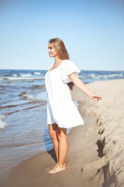 Okyanus sahilinde beyaz bir yaz elbisesi içinde kollarını açmış mutlu, güzel bir kadın..