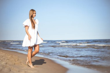 Mutlu gülümseyen güzel kadın beyaz bir yaz elbisesiyle okyanus sahilinde yürüyor..