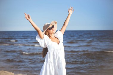 Mutlu sarışın kadın okyanus sahilinde beyaz bir elbise ve güneş gözlüğü ile el kaldırıyor..
