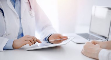 Doktor ve hasta klinikteki masada karşılıklı oturuyorlar. Tablet bilgisayar kullanan kadın doktorların ellerine odaklan, kapat. Tıp konsepti.