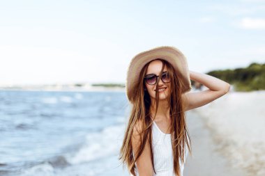 Okyanus sahilinde mutluluk içinde gülümseyen mutlu kadın şapka ve güneş gözlükleriyle poz veriyor. Tatil boyunca doğanın tadını çıkaran beyaz yaz elbiseli bir kadın modelin portresi.