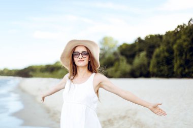 Okyanus sahilinde mutlu, mutlu, mutlu bir kadın şapkası, güneş gözlüğü ve açık elleriyle duruyor. Beyaz yaz elbisesi içinde çok kültürlü bir kadın modelin portresi seyahat sırasında doğanın tadını çıkarıyor.