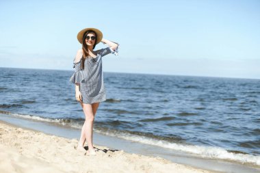 Okyanus sahilinde durup gülümserken kameraya bakan, moda şapkası ve güneş gözlüğü takan mutlu, genç esmer kadın. Keyifli tatil konsepti.