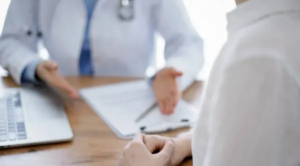 stock image Doctor and patient sitting and discussing something at wooden table while using a laptop computer. Focus is on patients hands. Medicine concept.