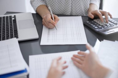 Kadın muhasebeci, hesap makinesi ve dizüstü bilgisayar kullanarak bir müşteriyle ya da meslektaşıyla vergileri sayıp tartışıyor. İş denetleme ekibi.