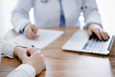 Doktor ve hasta oturmuş, dizüstü bilgisayar kullanırken ahşap masada bir şeyler tartışıyorlar. Hastaların ellerine odaklan. Tıp konsepti.