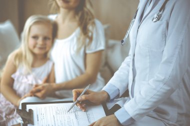 Doktor ve hasta. Pediyatrist, annesiyle birlikte küçük kızı muayene ederken pano kullanıyor. Sağlık muayenesinde mutlu beyaz çocuk. Tıp konsepti.