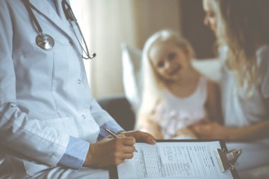 Doktor ve hasta. Pediyatrist, annesiyle birlikte küçük kızı muayene ederken pano kullanıyor. Sağlık muayenesinde mutlu beyaz çocuk. Tıp konsepti.