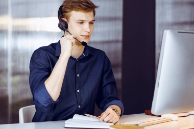 Genç sarışın iş adamı iş yerinde kulaklık ve bilgisayar kullanıyor. Girişim işi, başarı için çok çalışmak ve zamanın dışında çalışmak demektir..