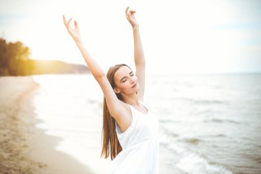 Mutlu gülümseyen kadın okyanus sahilinde elini kaldırarak mutlu mesut duruyor. Beyaz yaz elbisesi içinde çok kültürlü bir kadın modelin portresi seyahat tatilinde doğanın tadını çıkarıyor..