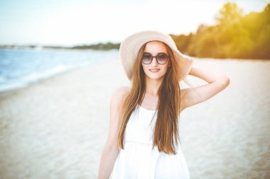 Okyanus sahilinde mutluluk içinde gülümseyen mutlu bir kadının portresi şapkası ve güneş gözlüğü ile duruyor. Beyaz yaz elbisesi içinde seyahat tatilinde doğanın tadını çıkaran bir kadın manken..