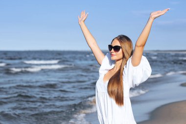 Mutlu sarışın kadın okyanus sahilinde beyaz bir elbise ve güneş gözlüğü ile el kaldırıyor..