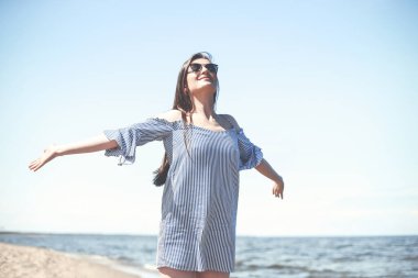 Okyanus sahilinde mutlu, mutlu ve mutlu bir kadın elleri açık duruyor. Yazlık elbiseli esmer bir kadın modelin portresi. Gezici tatiller sırasında doğanın tadını çıkarıyor..
