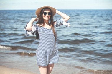 Okyanus sahilinde durup gülümserken kameraya bakan, moda şapkası ve güneş gözlüğü takan mutlu, genç esmer kadın. Keyifli tatil konsepti.