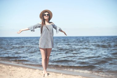 Okyanus sahilinde mutlu, mutlu ve mutlu bir kadın elleri açık duruyor. Güneş gözlüklü ve şapkalı esmer kadın model. Seyahat tatili boyunca doğanın tadını çıkarıyor..