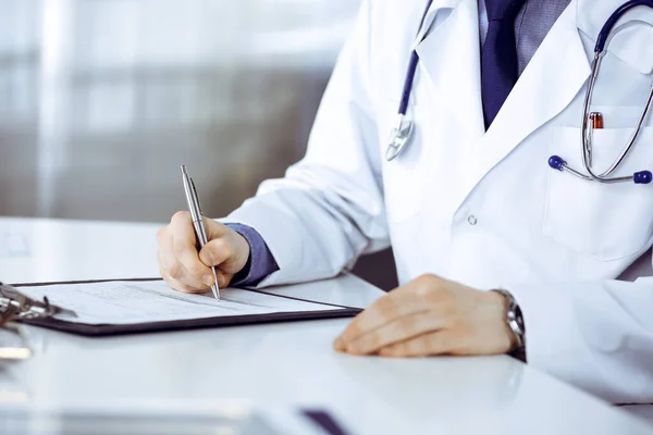 stock image Unknown male doctor sitting and working with clipboard of medication history record in clinic at his working place, close-up. Young physician at work. Perfect medical service, medicine concept.