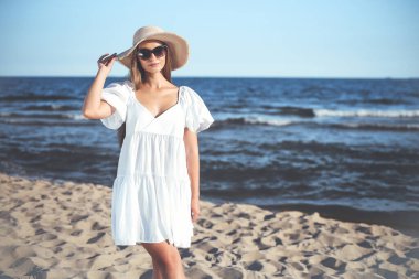 Mutlu sarışın kadın okyanus sahilinde güneş gözlüğü ve şapkasıyla poz veriyor. Akşam güneşi.