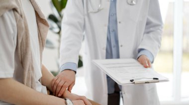 Doktor ve hasta klinikte. Bir yandan dostane bir doktor genç bir kadını muayene ederken diğer yandan tıbbi kayıtlarla dolu bir pano tutuyor. Tıp konsepti.