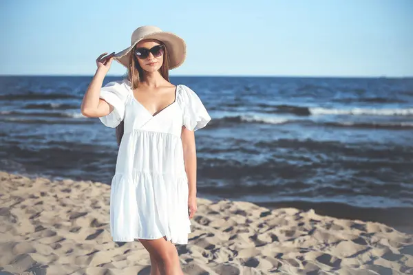 Happy blonde woman is posing on the ocean beach with sunglasses and a hat. Evening sun.