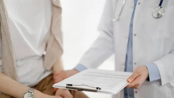 Doctor and patient discussing health exam results. Friendly physician reassuring a young woman by one hand while keeping clipboard with medical papers in another. Medicine concept.