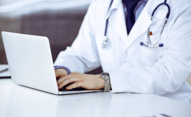 Bilinmeyen bir erkek doktor klinikte dizüstü bilgisayarla yakın planda çalışıyor. İşteki genç doktor. Mükemmel sağlık hizmeti, tıp konsepti.