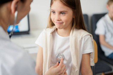 Doktor ve çocuk hasta klinikte. Beyaz önlüklü bir doktor steteskopla gülümseyen genç bir kızı muayene ediyor. İlaç, terapi konsepti.